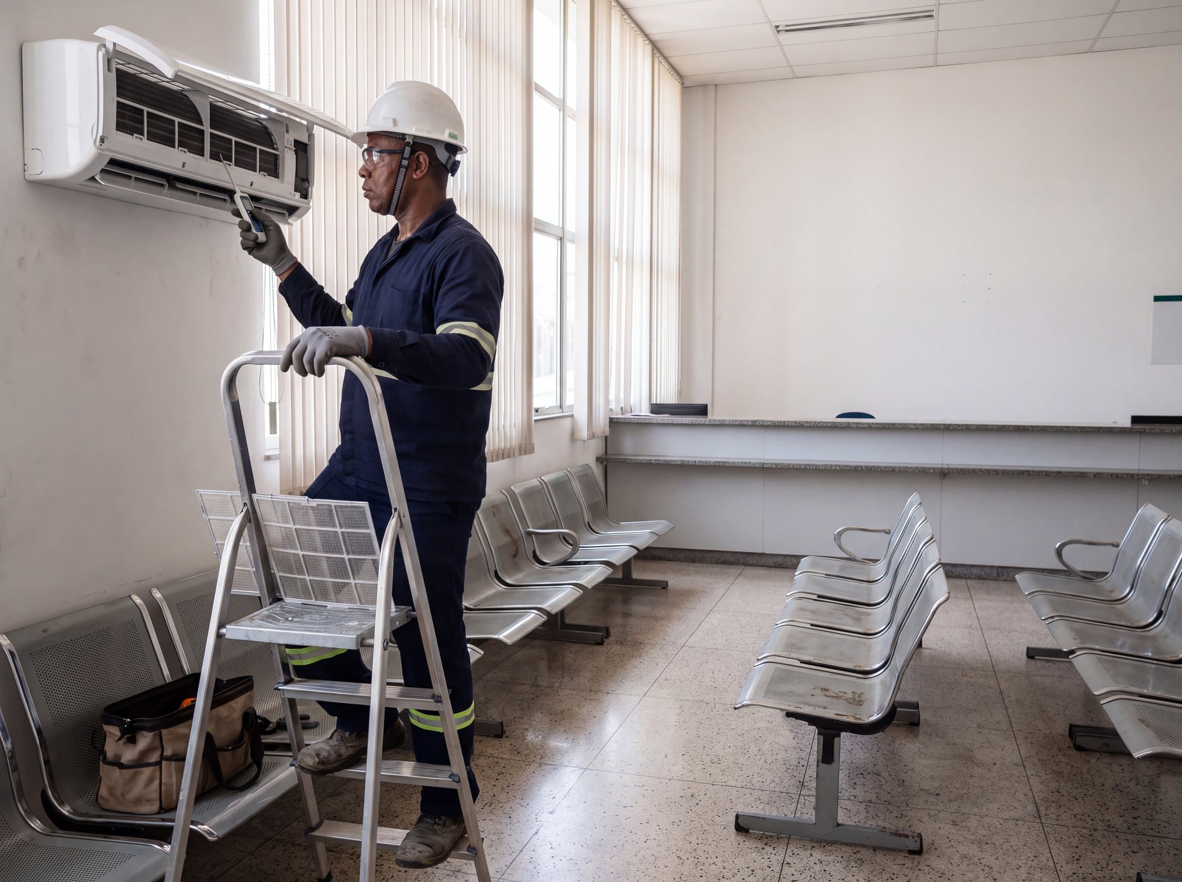Técnico de capacete, óculos e luvas sobre uma escada medindo a evaporadora aberta na parede de uma sala de atendimento com cadeiras de espera vazias