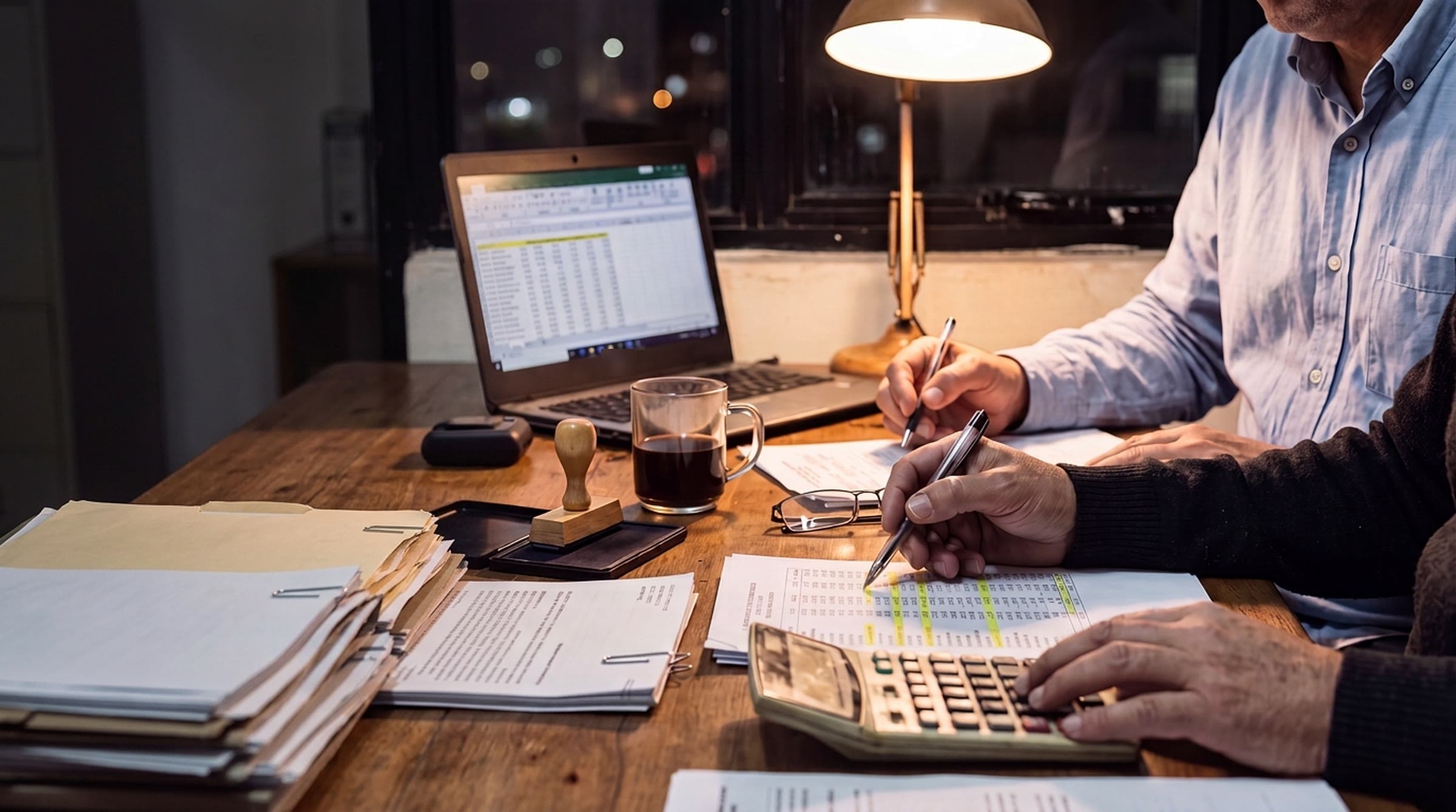 Mesa de escritório à noite sob luminária: duas pessoas conferem planilhas impressas com caneta e calculadora, ao lado de um carimbo, uma xícara de café e o notebook com a planilha aberta