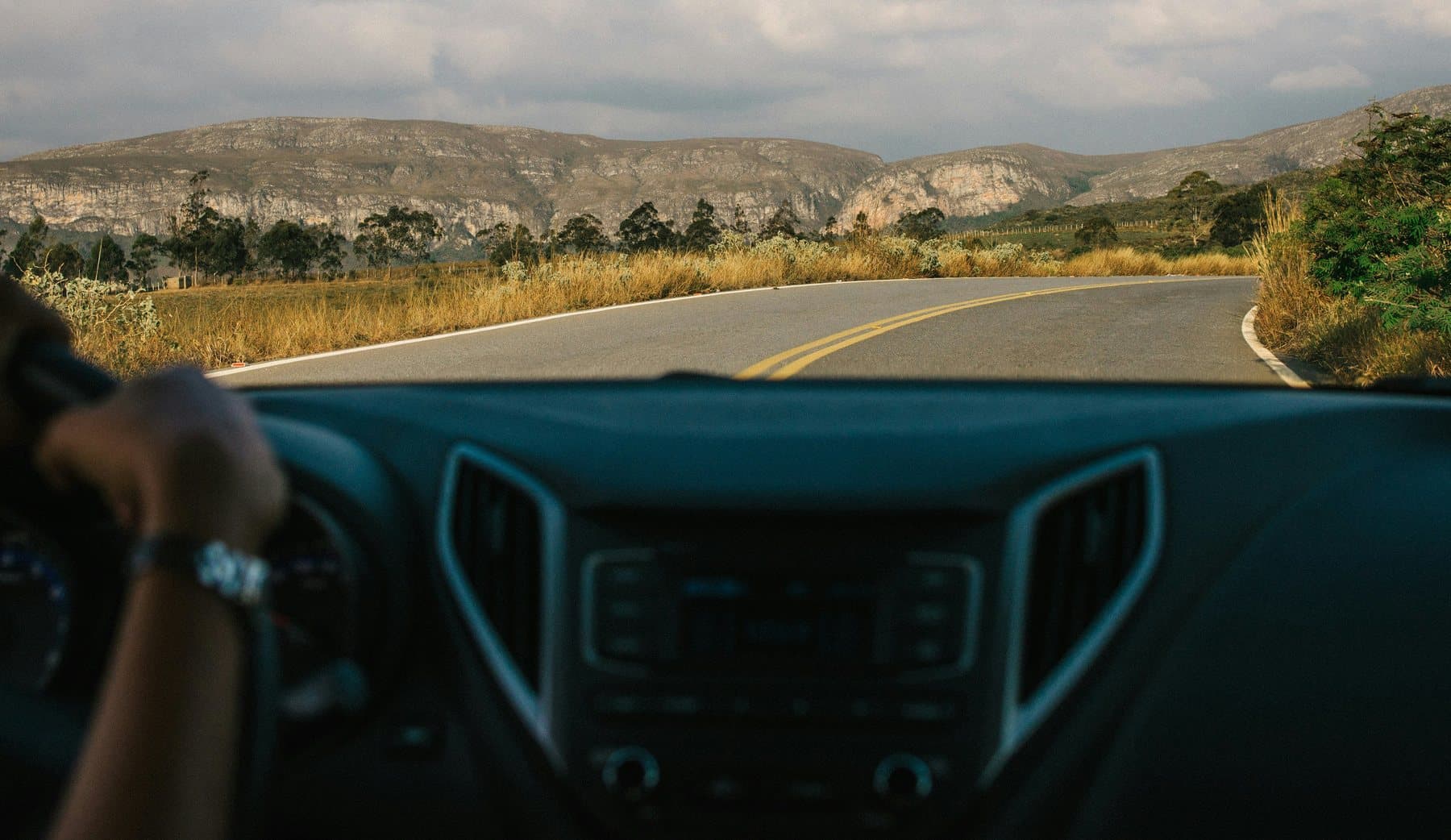 Vista do para-brisa de um carro numa estrada de serra em Minas Gerais, com o paredão de pedra da Serra do Cipó no horizonte e capim dourado nas margens